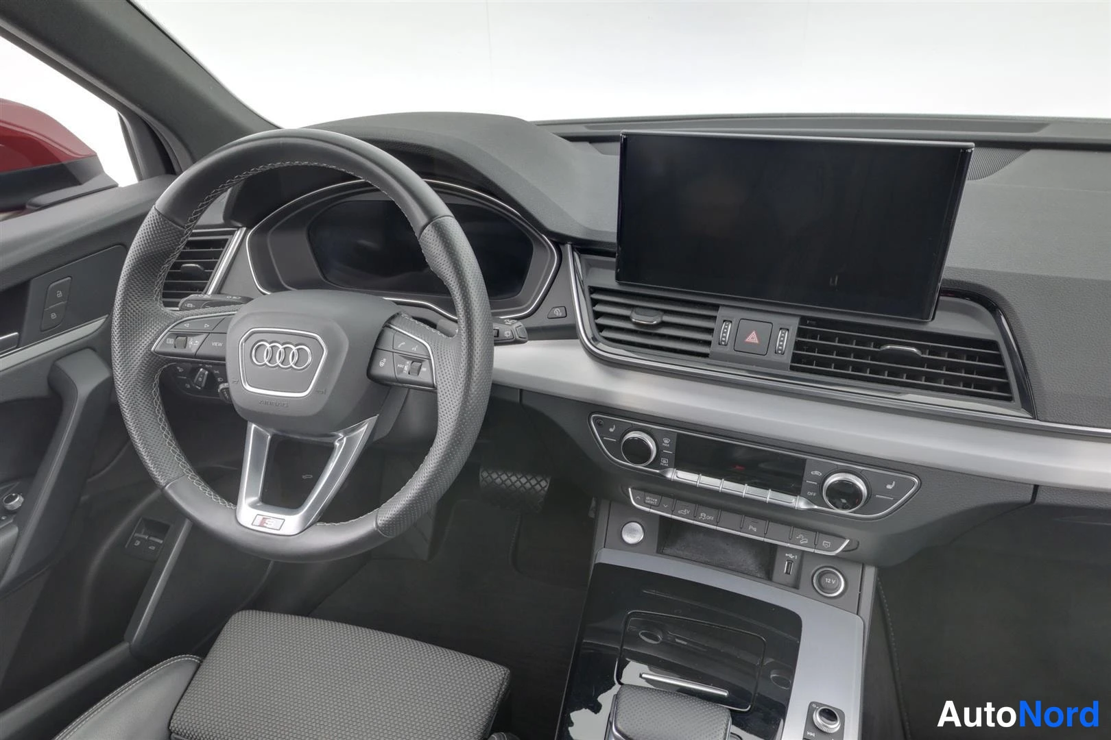 Edestä: Lähikuva etupaneelista: Audi-merkkinen ohjauspyörä, kolmiot Suuri keskinäinen näyttö ja keskikonsolin säätimet. Kojelaudan musta pinta ja ohjauspyörän tekstiili
