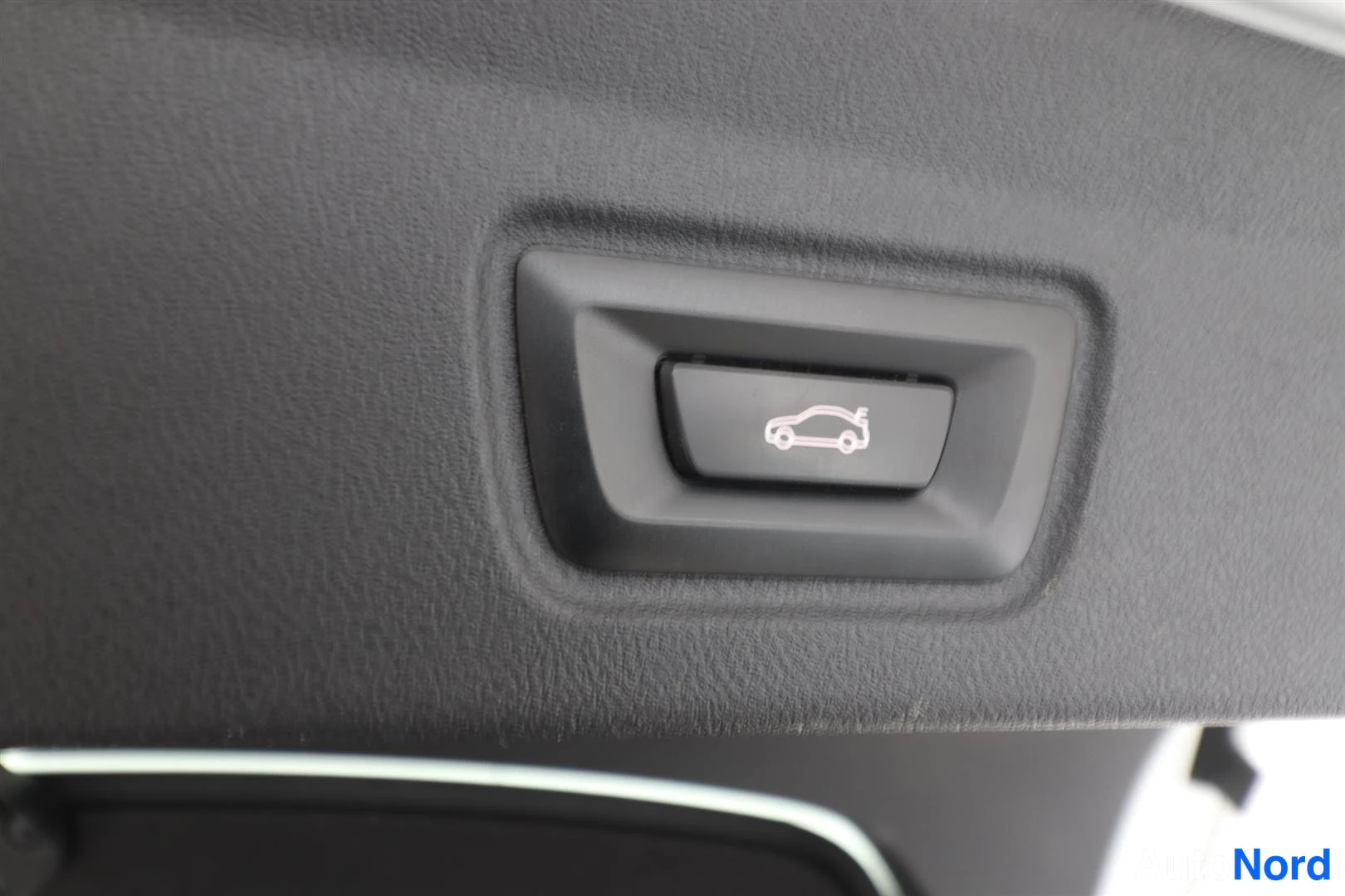 Yksityiskohta: Close-up of a black rectangular push-button switch mounted on a textured dark interior panel. The button has a white line-drawing icon depicting the side profil