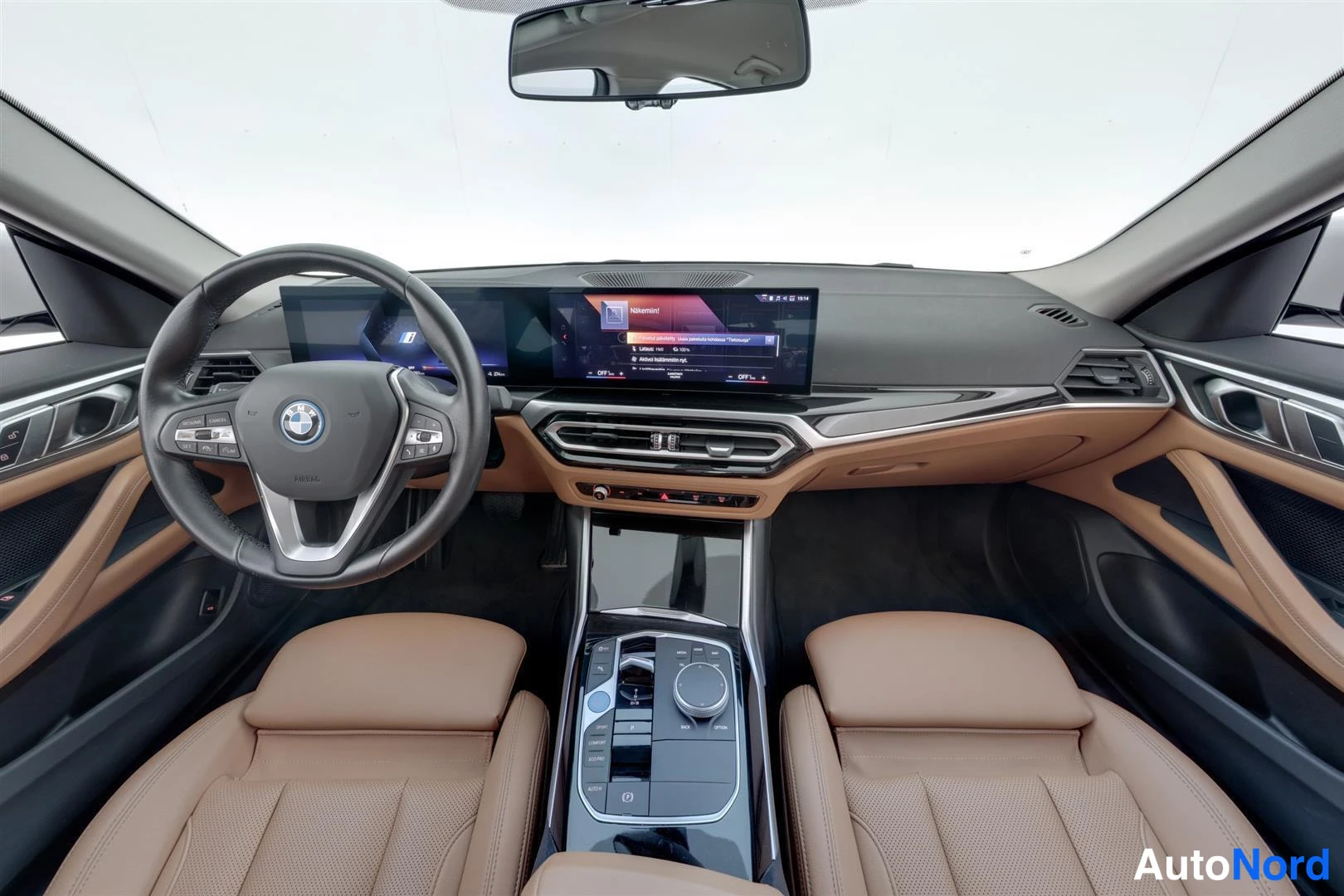 Muu: Sisätilanäkymä takaa eteenpäin: BMW-ohjauspyörä, pitkä kaareva infotainment-näyttö, ruskeat nahkaistuimet ja keskikonsoli, jonka iDrive-ohjain näkyy.