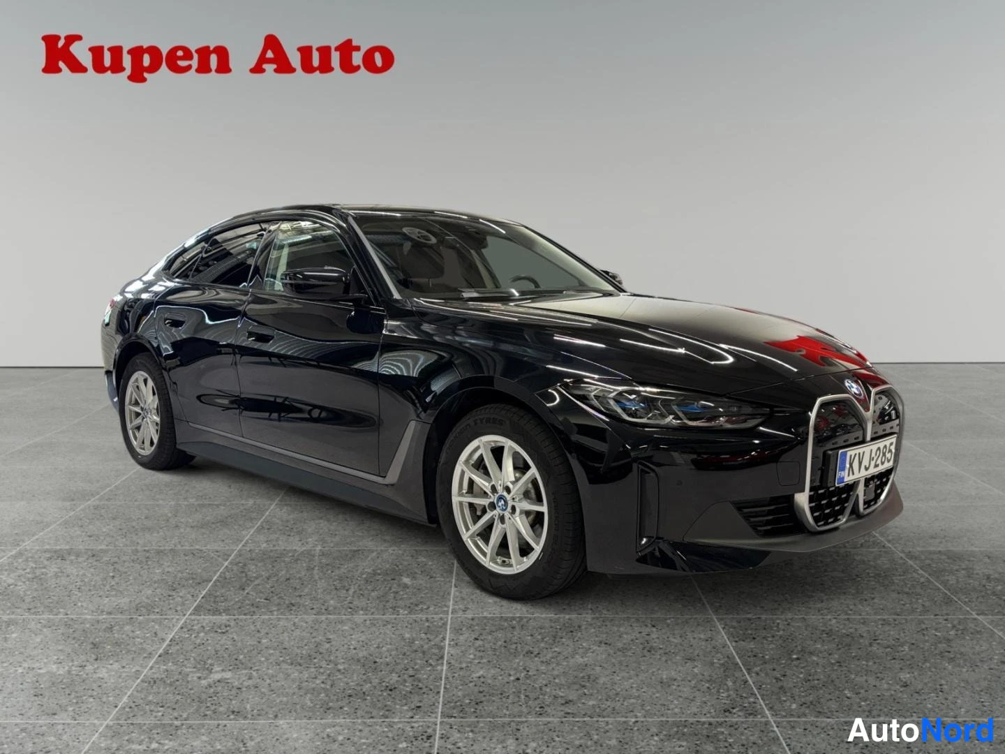 Etuviisto, nokka oikealle: Mustan BMW i4 -sedanin etukulmanäkymä oikealta, rekisterikilpi, valokuvattuna autosalon harmaassa ympäristössä.