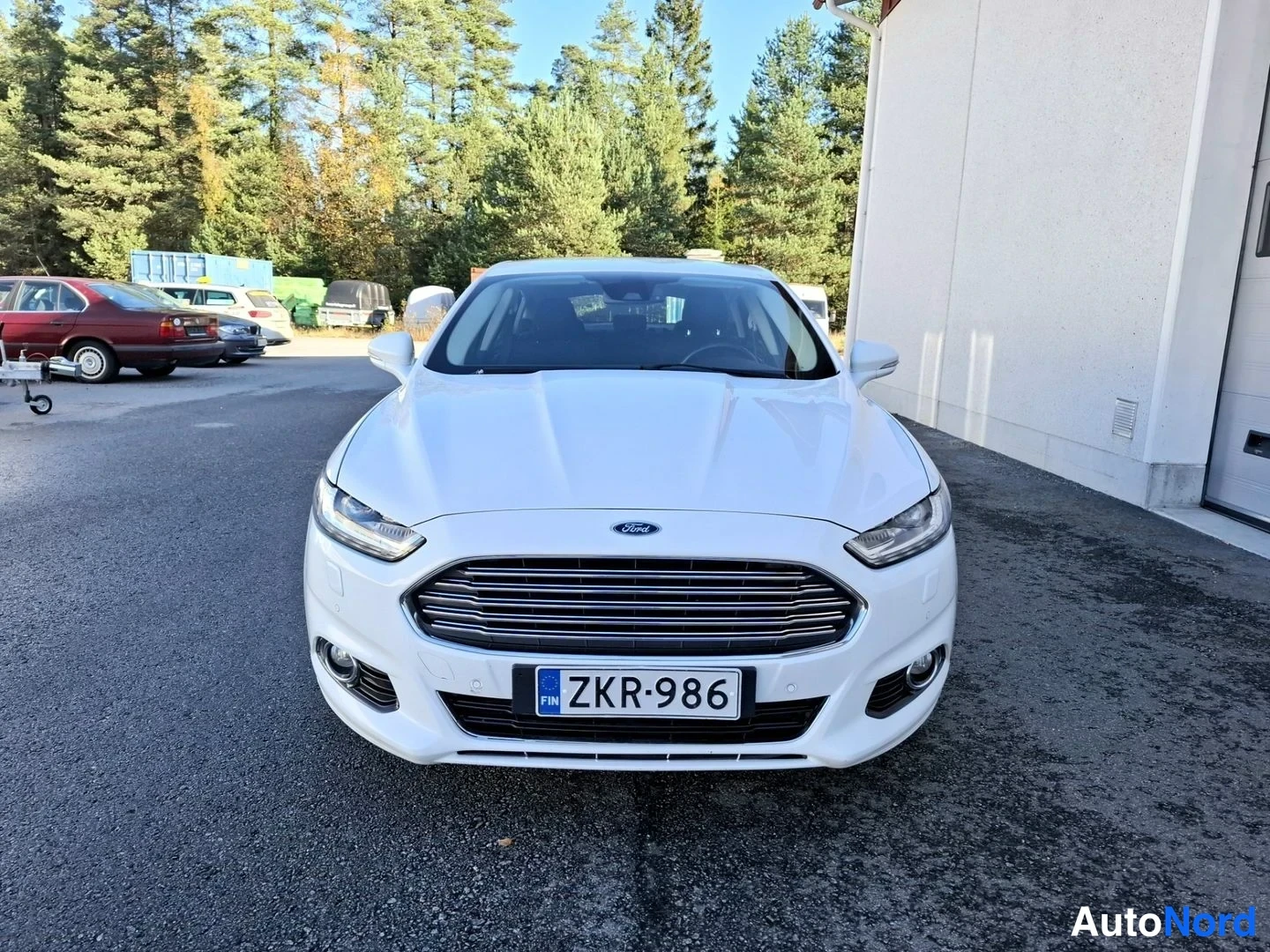 Edestä: Valkoinen Ford Mondeo edestäpäin parkkeerattuna rakennuksen vieressä, rekisteritunnus.