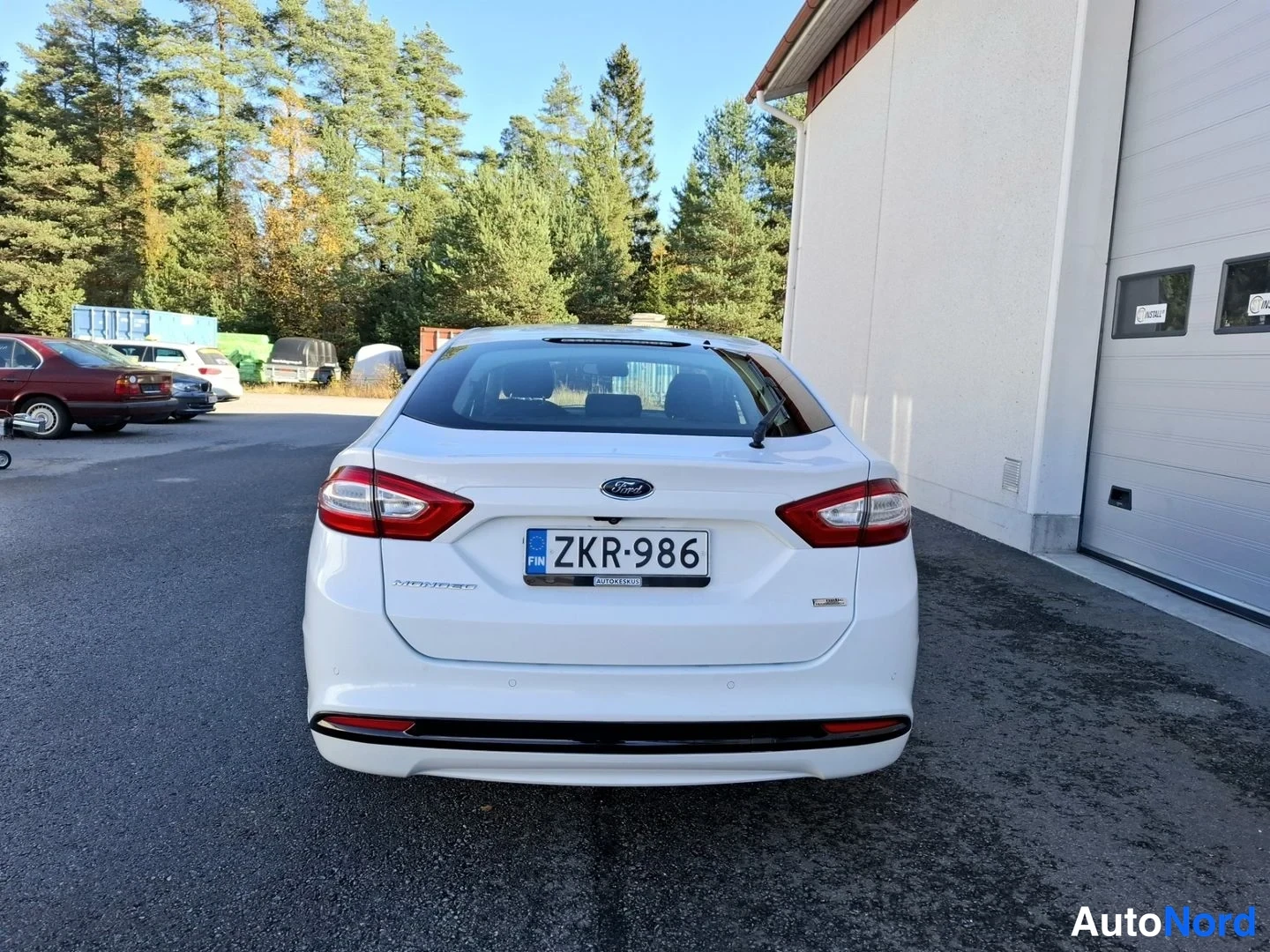 Takaa: Valkoisen Ford Mondeon takapää takaa kuvattuna, rekisteritunnus, taustalla metsää ja rakennus.