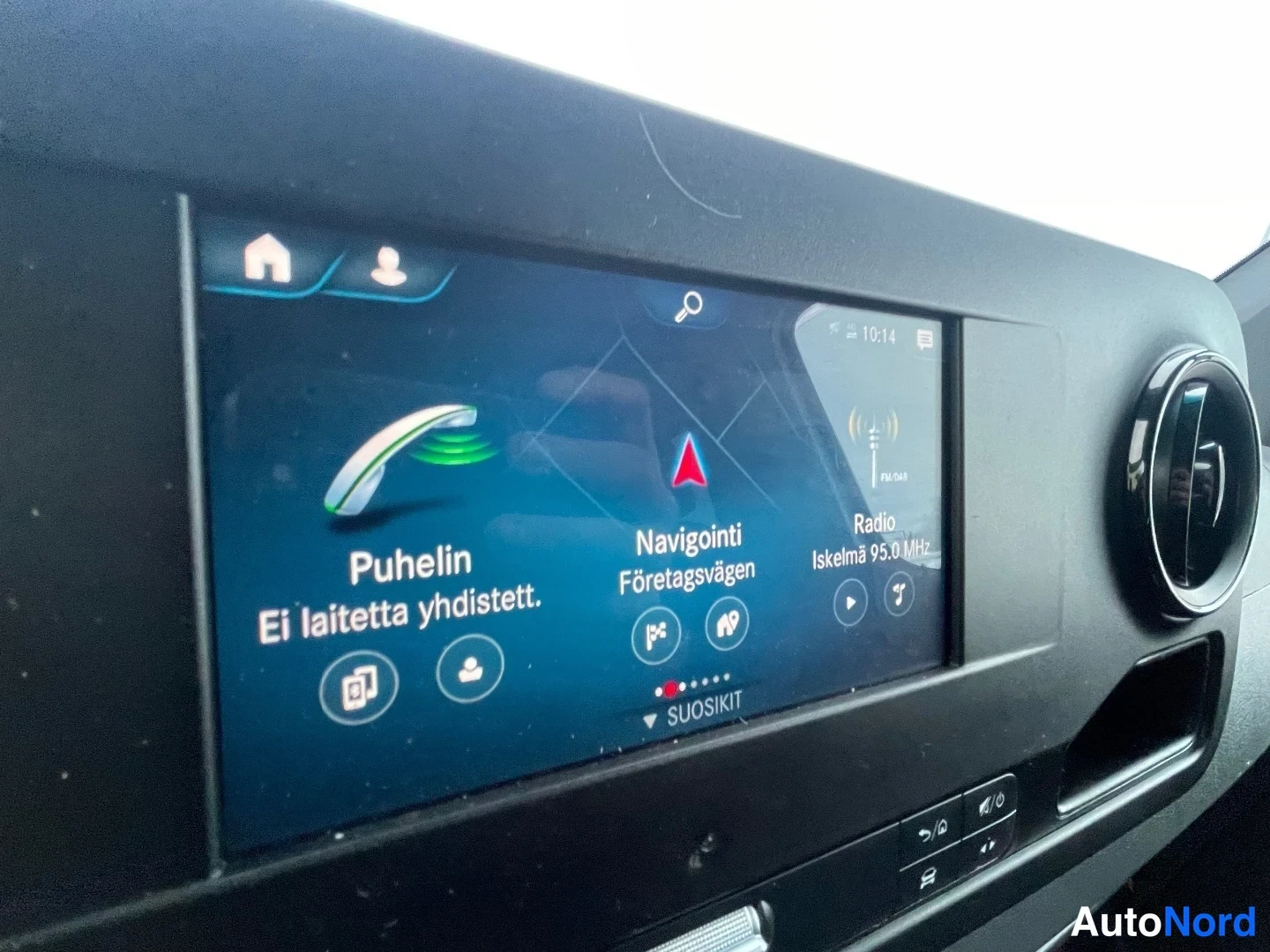 Etuviisto, nokka vasemmalle: Sisätarkka kuva auton kojelaudan infotainment-näytöstä, jossa näkyy valikko: Puhelin (Ei laitetta yhdistett.), Navigointi Företagsvägen ja Radio Iskelm