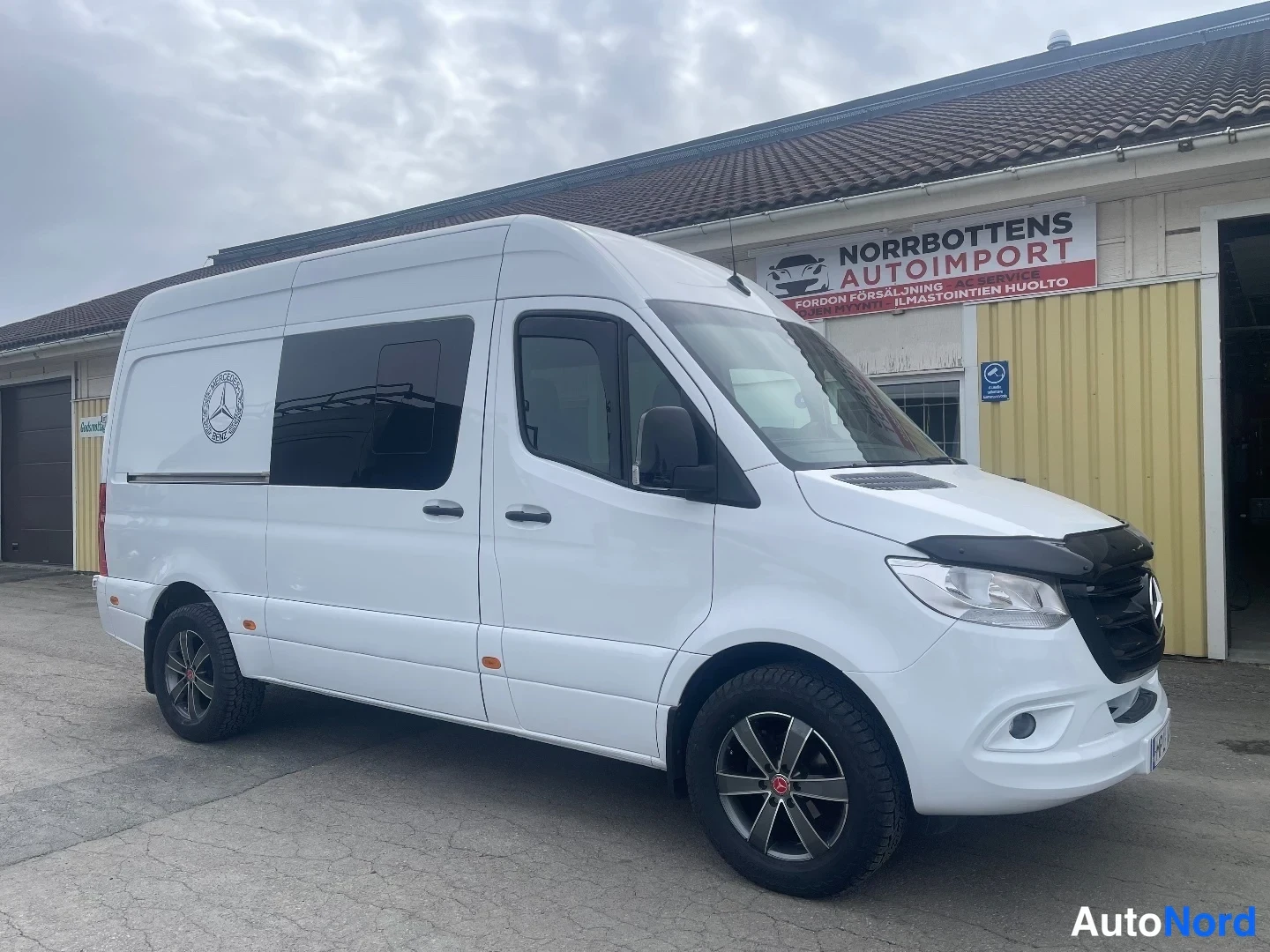 Etuviisto, nokka oikealle: Valkoinen Mercedes-Benz Sprinter etuviisto vasemmalta liiketalon edustalla, konepellin eteen asennettu musta hupisuoja ja kevytmetallivanteet.