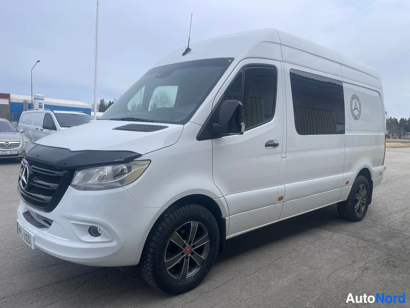 Etuviisto, nokka vasemmalle: Valkoinen Mercedes-Benz Sprinter -pakettiauto etuviisto vasemmalta, tumma etusäleikkö ja tummat kevytmetallivanteet.