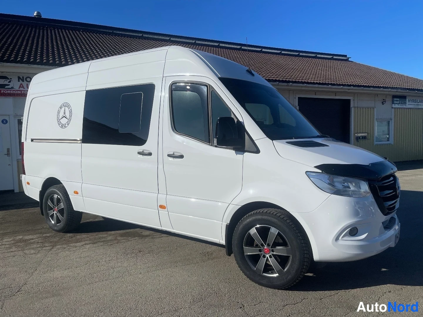 Etuviisto, nokka oikealle: Valkoinen Mercedes-Benz Sprinter -pakettiauto kolmannelne asema oikealta edestä, korkealla katolla, merkki kyljessä.