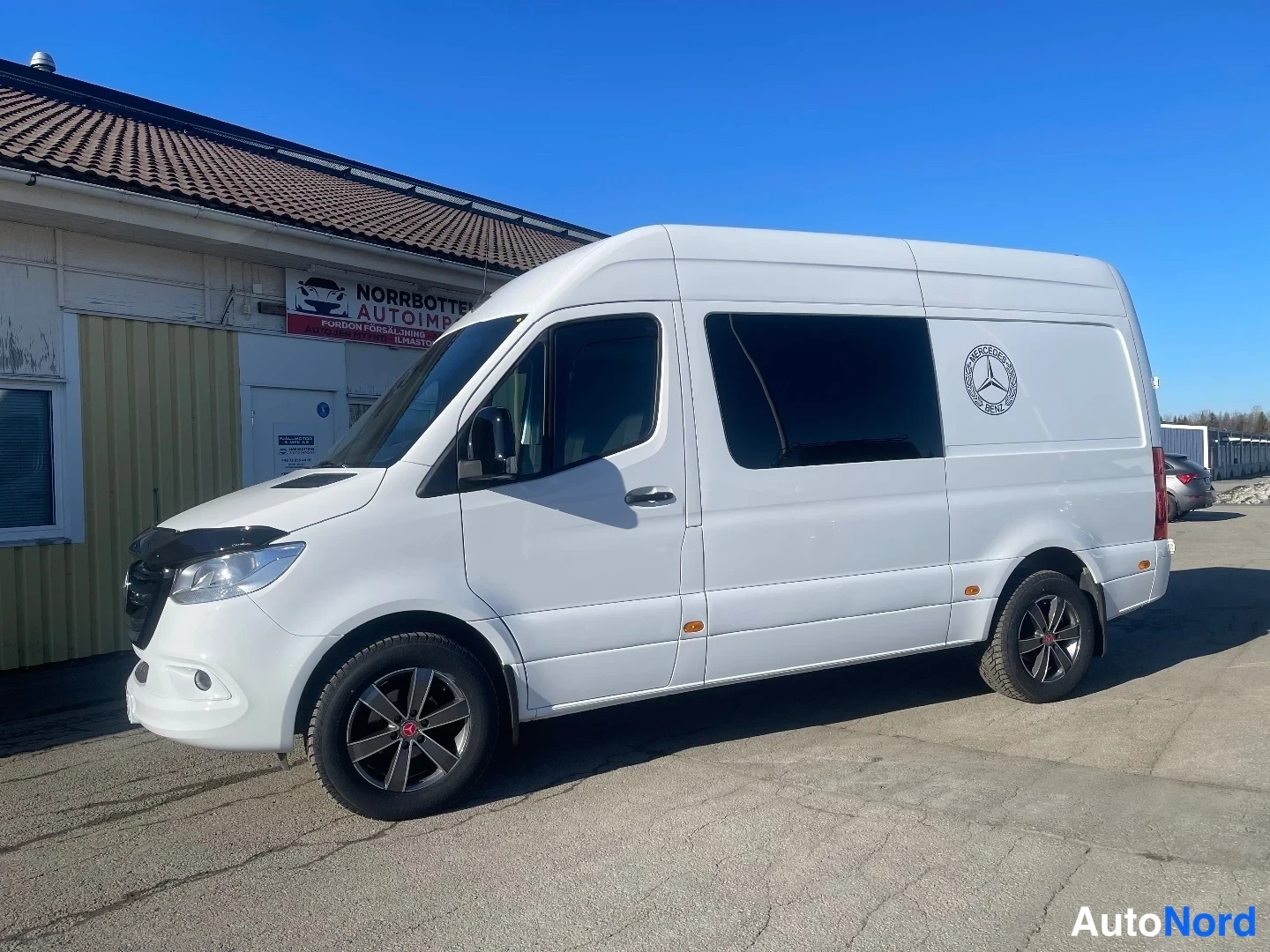 Etuviisto, nokka vasemmalle: Valkoinen Mercedes-Benz Sprinter -pakettiauto kolmannelne asema vasemmalta edestä, kori korkea ja markiisit ikkunoissa.