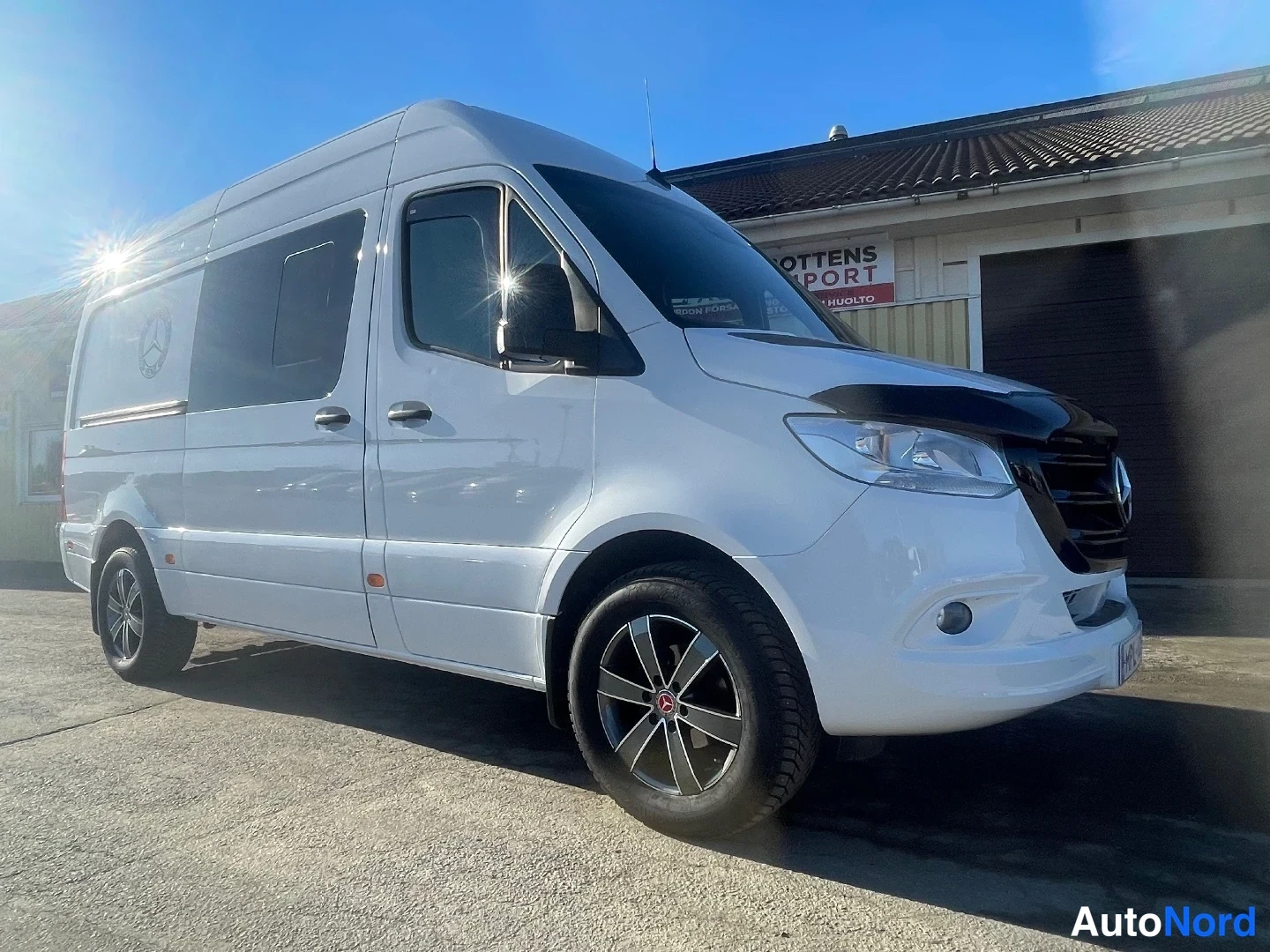 Etuviisto, nokka oikealle: Valkoisen Mercedes-Benz Sprinter -pakettiauton etukulma, konepelti ja kylki auringonvalossa