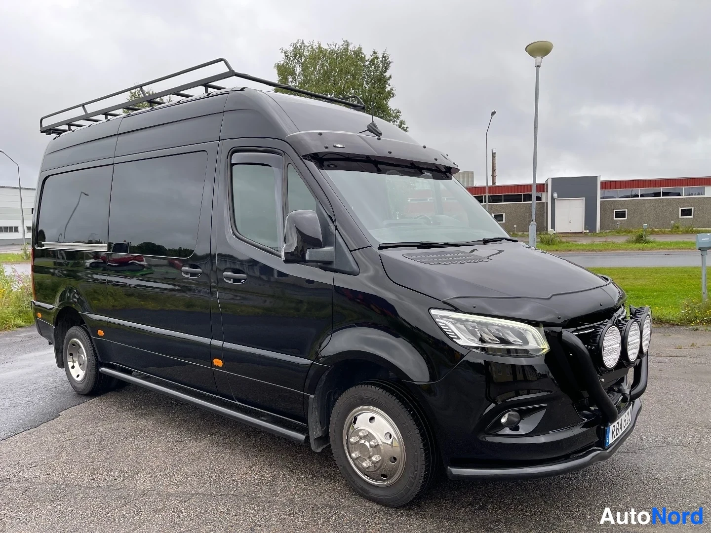 Etuviisto, nokka oikealle: Musta Mercedes-Benz Sprinter -pakettiauto kolmiosta etuviistöstä, kattoteline ja lisävalot.