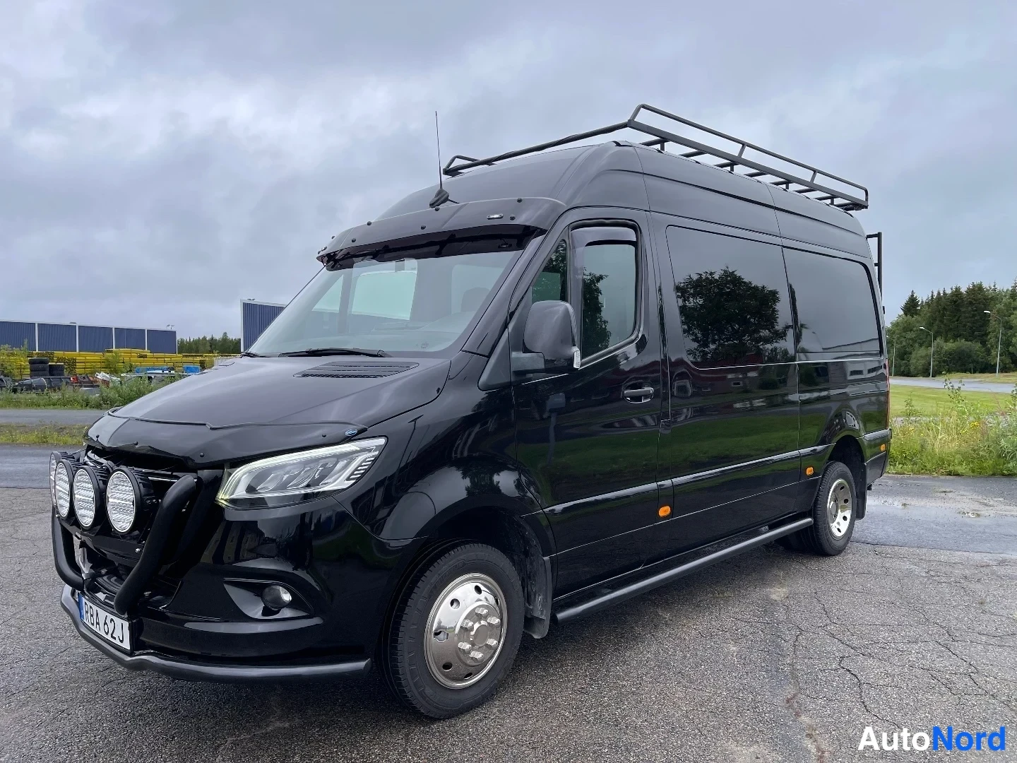 Etuviisto, nokka vasemmalle: Musta Mercedes-Benz Sprinter -pakettiauto etuviistöstä vasemmalta, kattoteline ja lisävalot.