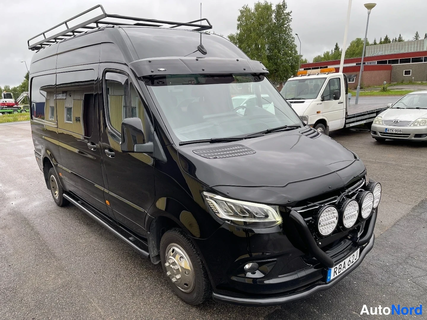 Etuviisto, nokka oikealle: Musta Mercedes-Benz Sprinter -pakettiauto edestä oikealta kuvattuna; keulakaariin asennettuna neljä lisävaloa ja katolla tavarateline.