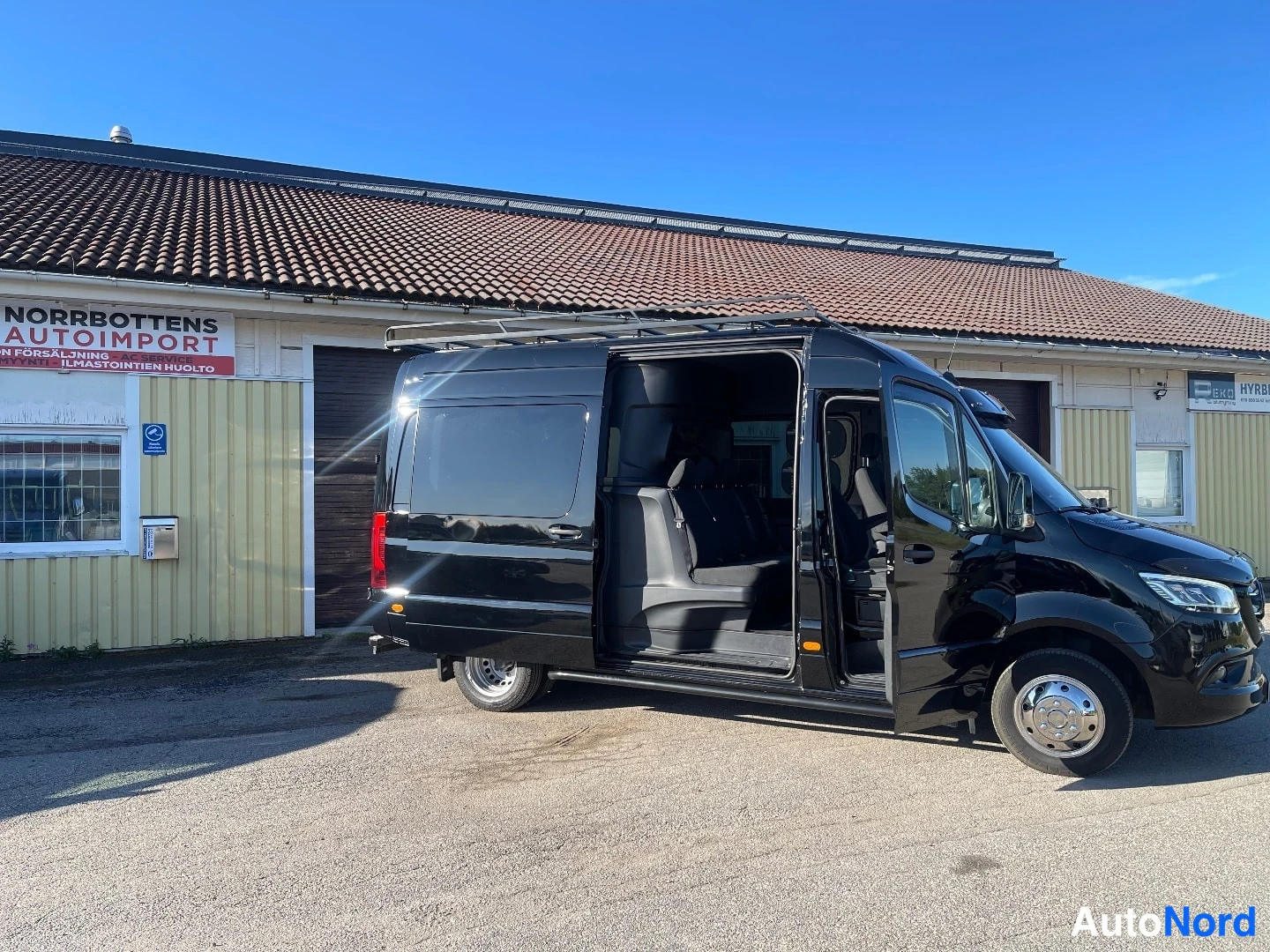 Etuviisto, nokka oikealle: Musta Mercedes-Benz Sprinter -pakettiauto sivusta oikealta kuvattuna; liukuova ja etuovi auki ja sisätiloissa näkyy istuimia.