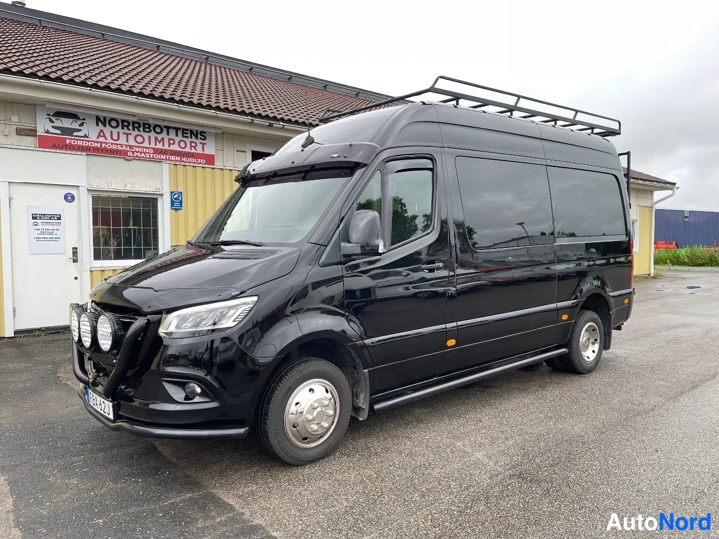 Etuviisto, nokka vasemmalle: Musta Mercedes-Benz Sprinter -pakettiauto etuviistöstä vasemmalta huoltorakennuksen edessä, kattoteline ja lisävalot.