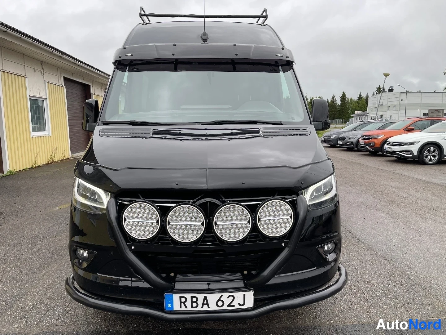 Edestä: Edessäpäin kuvattu musta pakettiauto, jonka keulassa neljä lisävaloa ja kattoteline; rekisterikilpi RBA 62J.