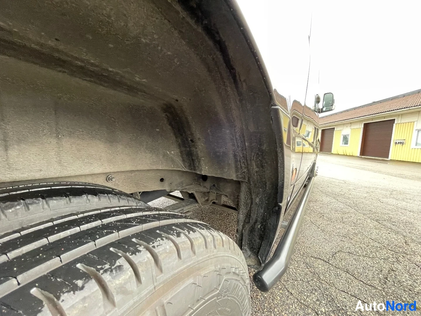 Yksityiskohta: Lähikuva auton etupyörästä ja likaisesta konepellin alta avattuna; sivuoven kynnyslista näkyy oikealla.