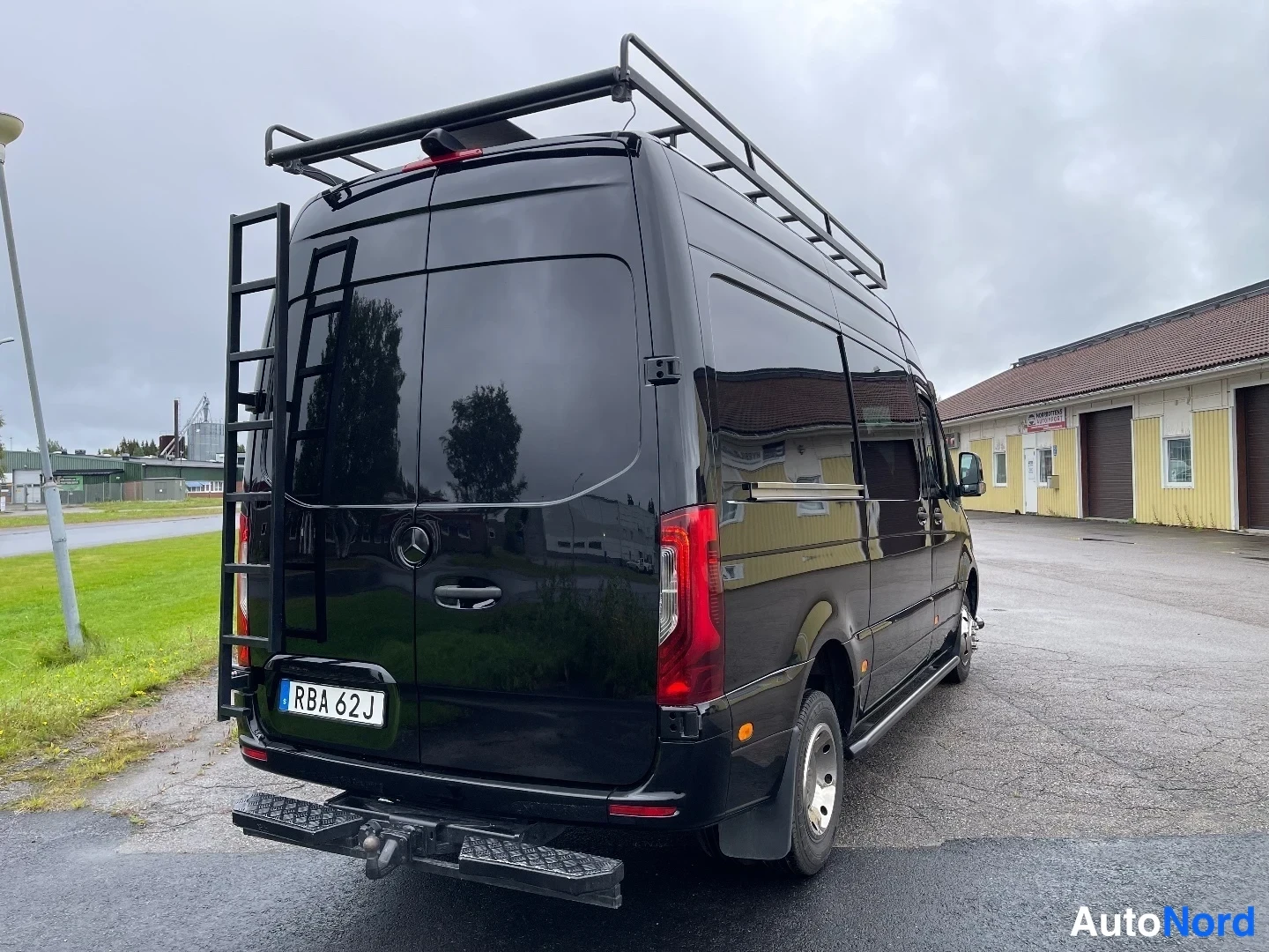 Takaviisto: Mustan Mercedes-Benz Sprinter -pakettiauton takaosa ja oikea kylki, katolla kaariteline, perässä tikkaat ja vetokoukku.