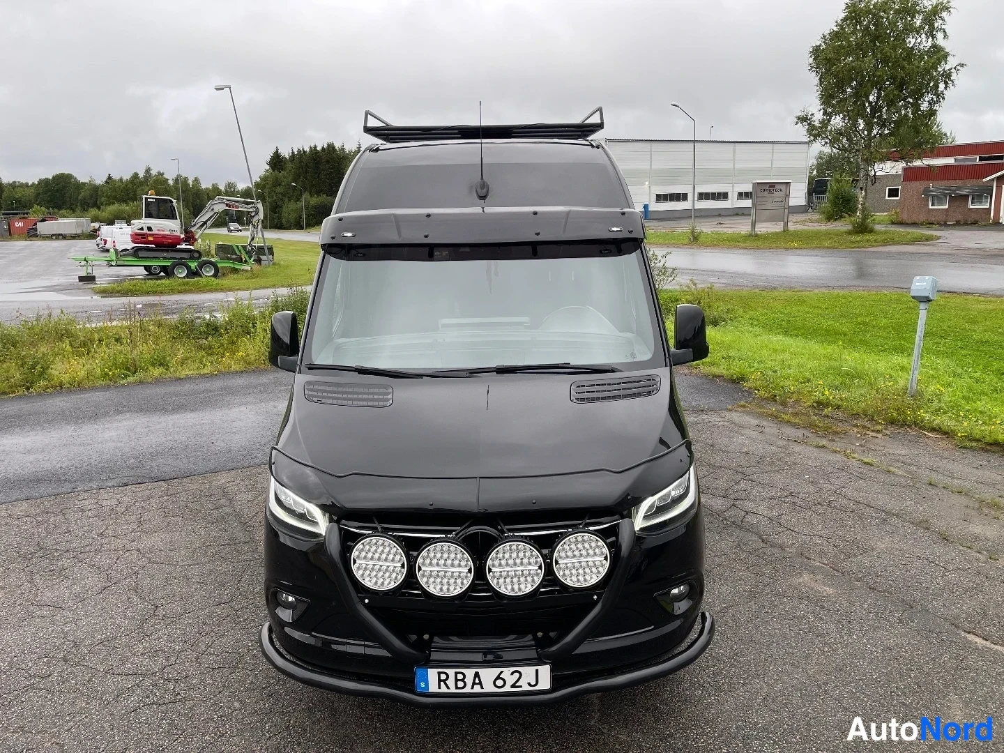 Edestä: Mustan Mercedes-Benz Sprinter -pakettiauton etunäkymä, keulassa neljä suurta pyöreää lisävaloa, katolla kaariteline.