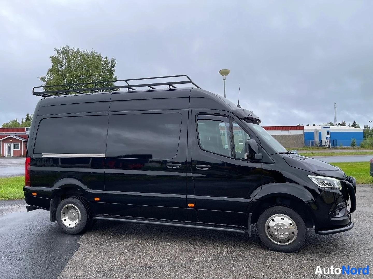 Etuviisto, nokka oikealle: Mustan Mercedes-Benz Sprinter -pakettiauton vasen kylki ja etuosa, katolla kaariteline, tummat ikkunat.
