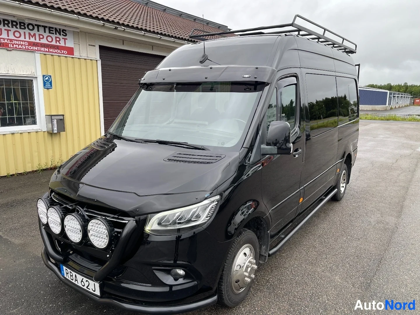 Etuviisto, nokka vasemmalle: Mustan Mercedes-Benz Sprinter -pakettiauton etuosa ja oikea kylki autotallin edessä, keulassa lisävalot, katolla kaariteline.