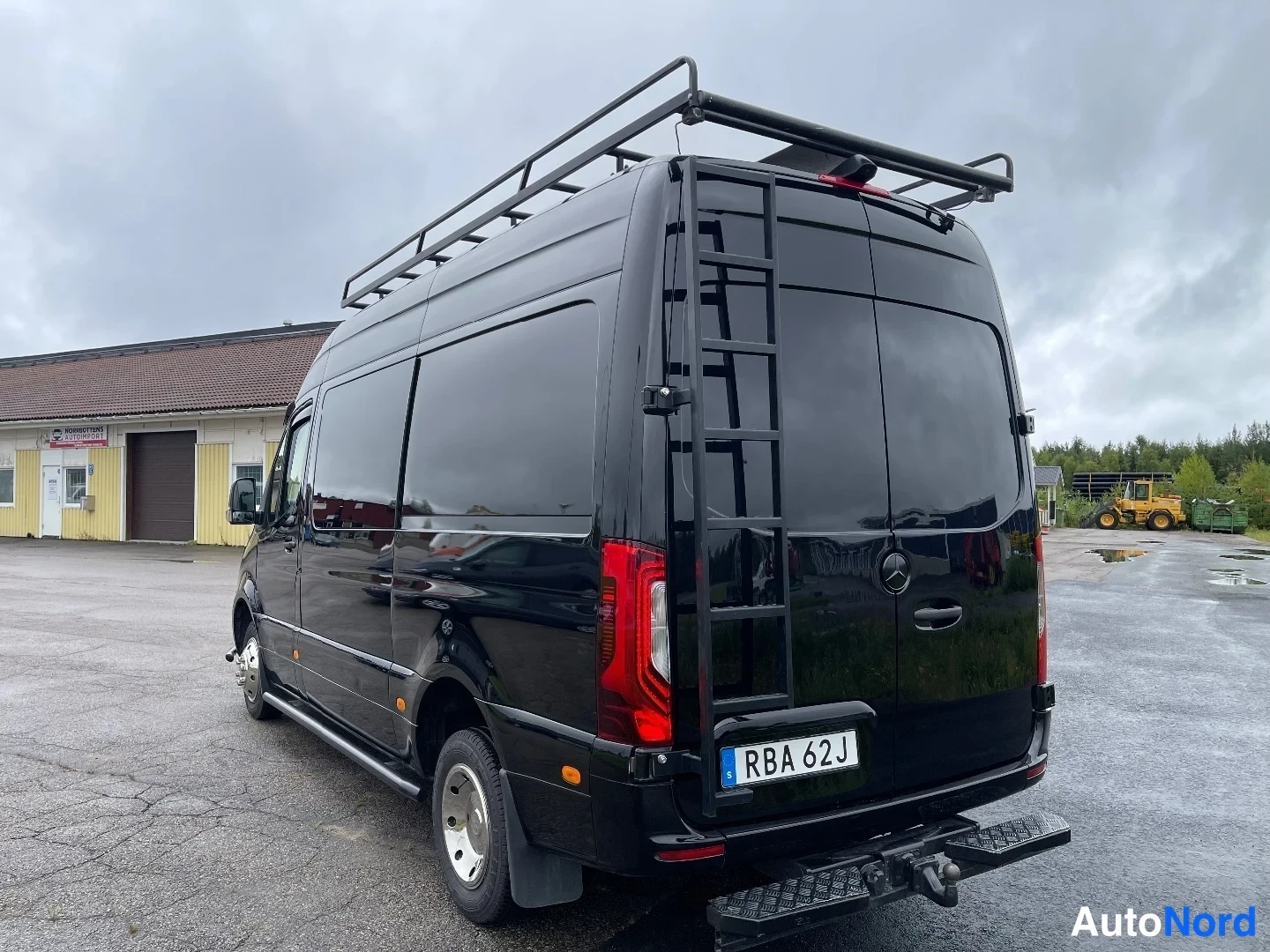 Takaviisto: Musta Mercedes-Benz Sprinter -pakettiauto takaa vasemmalta kuvattuna; takaovessa tikkaita ja katolla tavarateline, perässä vetokoulu.