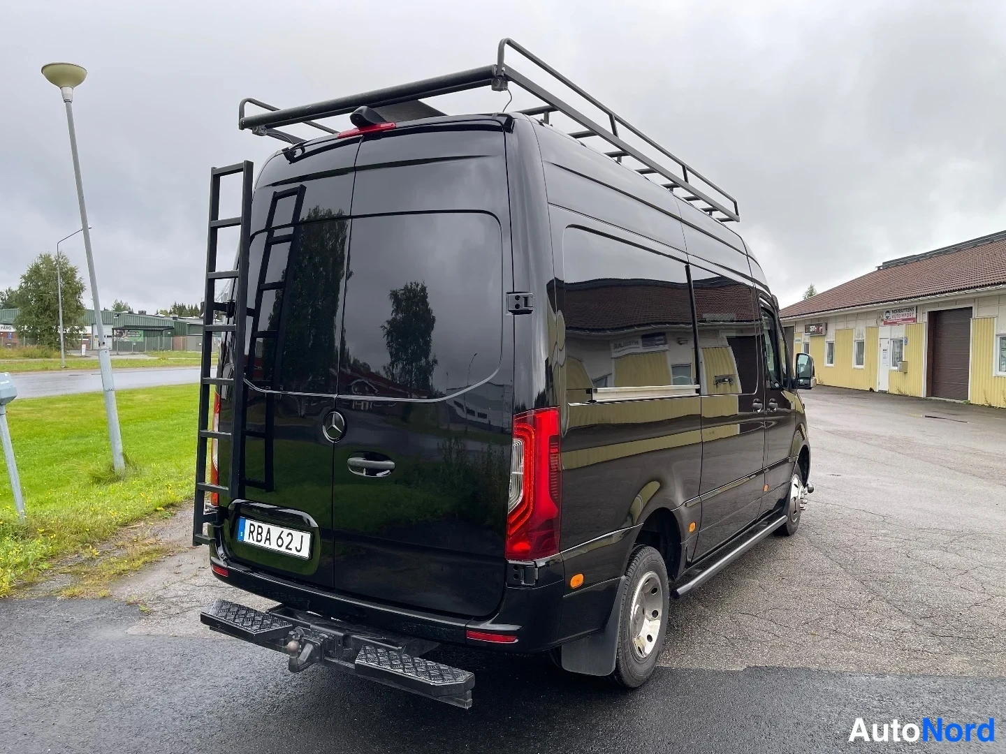 Takaviisto: Musta Mercedes-Benz Sprinter -pakettiauto takaa oikealta kuvattuna; takaovessa tikkaita ja katolla tavarateline.