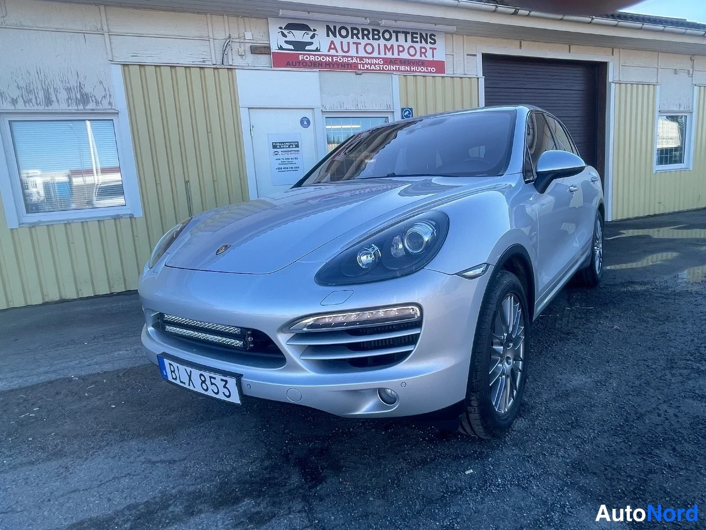 Etuviisto, nokka vasemmalle: Porsche Cayenne -farmarikorinen SUV hopeanvärisenä edestä vasemmalta kolmivartalokuvassa tontin pihalla.