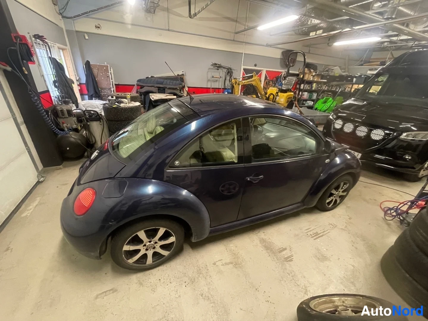 Takaviisto: Tummansininen Volkswagen New Beetle -henkilöauto takaviistosta varastotilassa, takavalo ja oikea kylki näkyvissä.