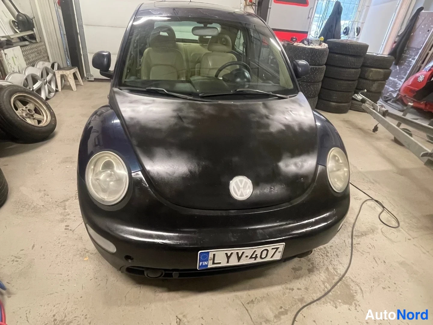 Edestä: Volkswagen New Beetlein keula edestä päin hallissa, rekisterikilpi, konepelti pölyinen.