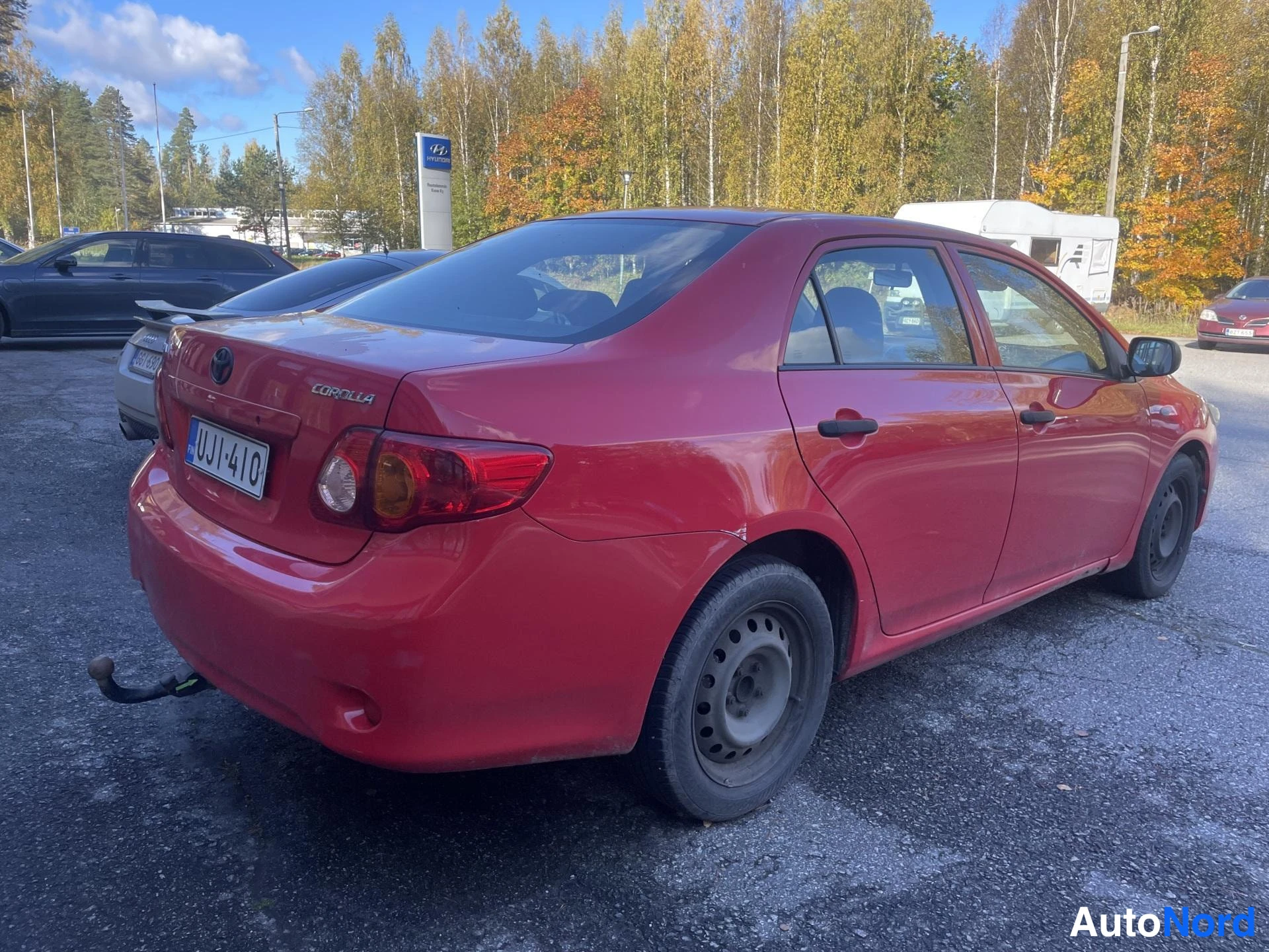 Takaviisto: Punaisen sedanin takaosa ja oikea sivu kuvattuna parkkipaikalla, taustalla syksyisiä puita ja muita autoja.