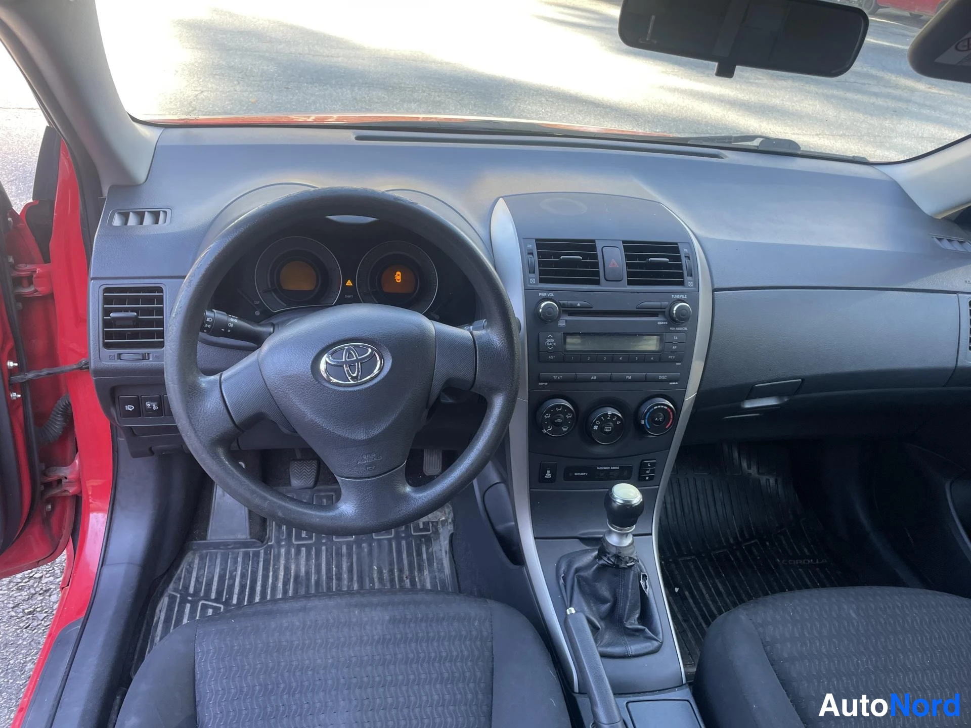 Edestä: Toyota Corollan kojelauta suoraan edestä: Toyota-merkkinen ohjauspyörä, radio ja manuaalivaihteen keppi.
