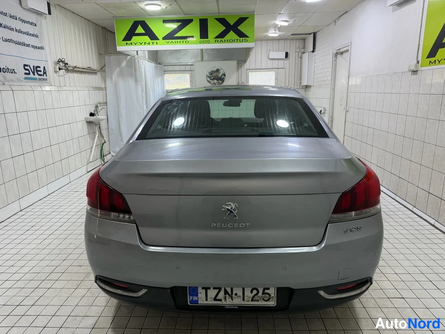 Takaa: Auton takaosa suoraan takaa, rekisterikilpi ja Peugeot-merkintä konepellin takana, laatoitetussa sisätilassa.