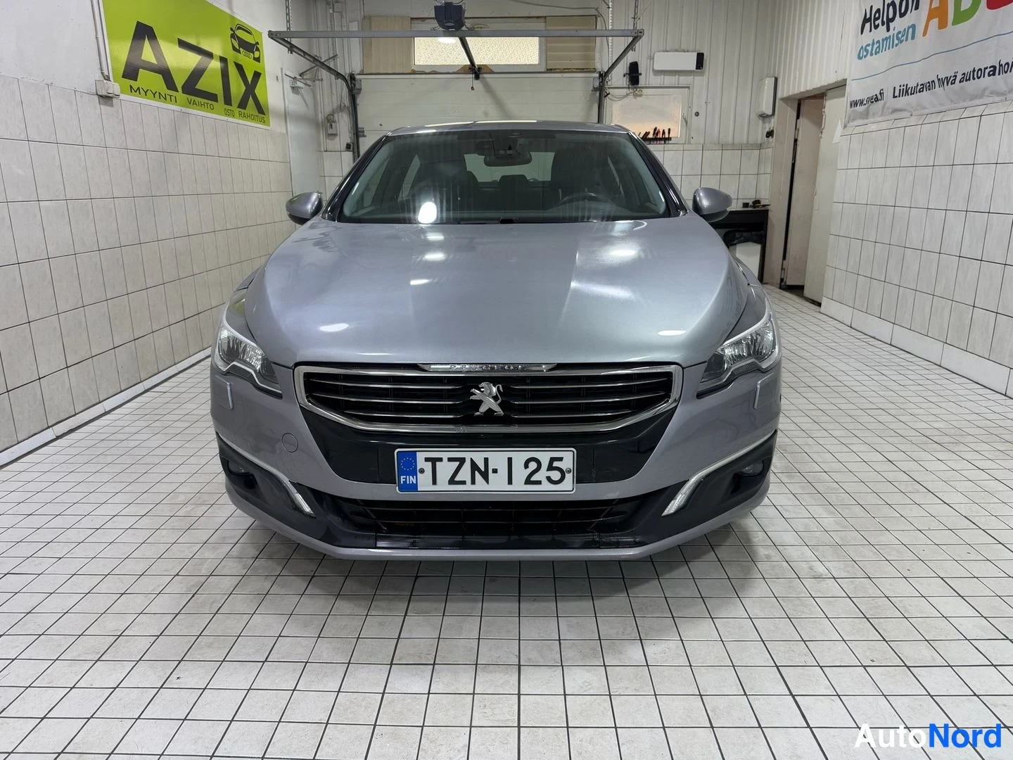 Edestä: Auton keula suoraan edestä; hopeinen Peugeot-sedan, rekisterikilpi, AZIX-kyltti taustalla.