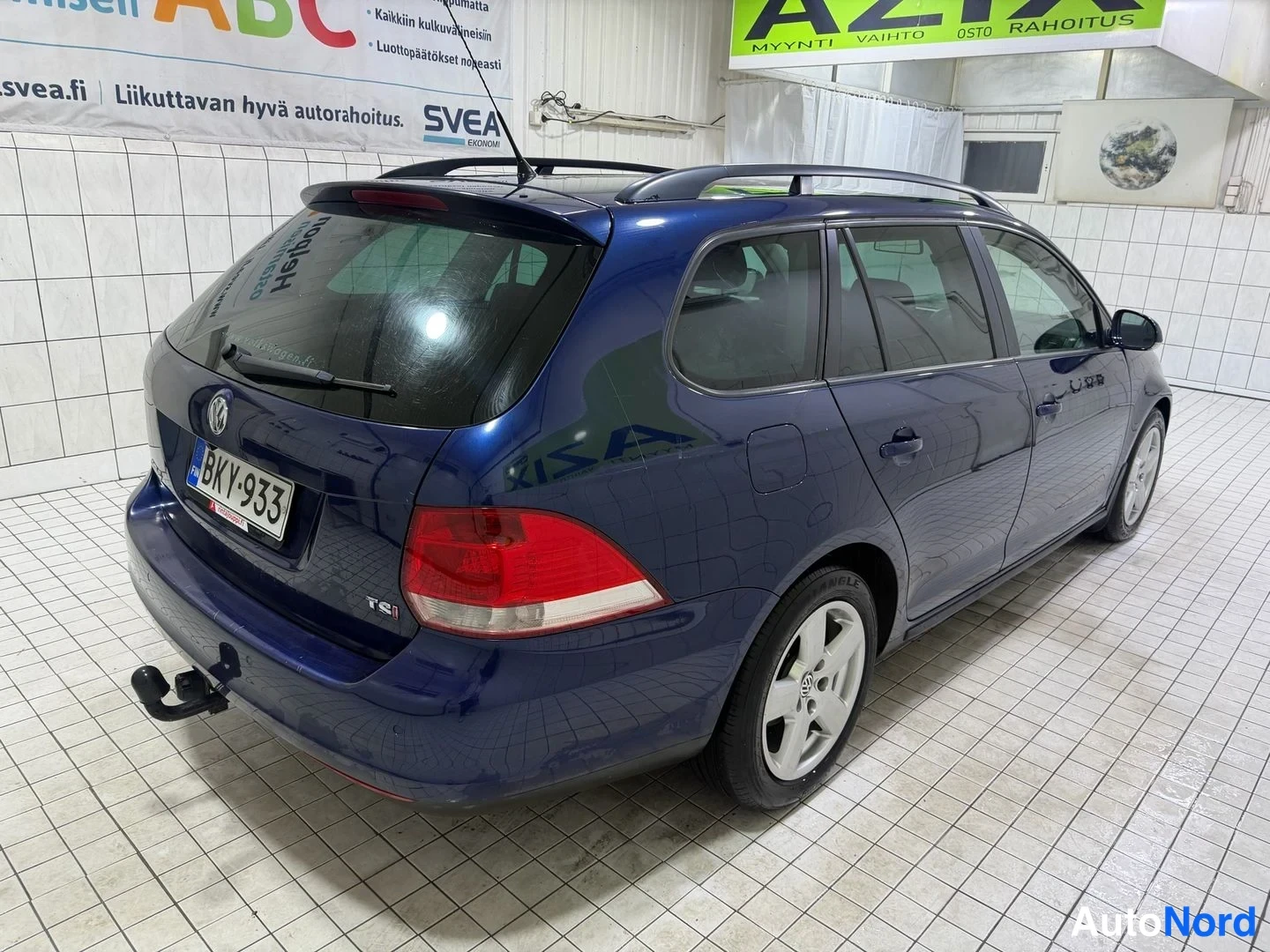 Takaviisto: Volkswagen Golf Variant -farmariauto takaa kulmasta valokuvattuna sisätiloissa; tummansininen koripinta, kattokaiteet ja vetokoukku näkyvät.
