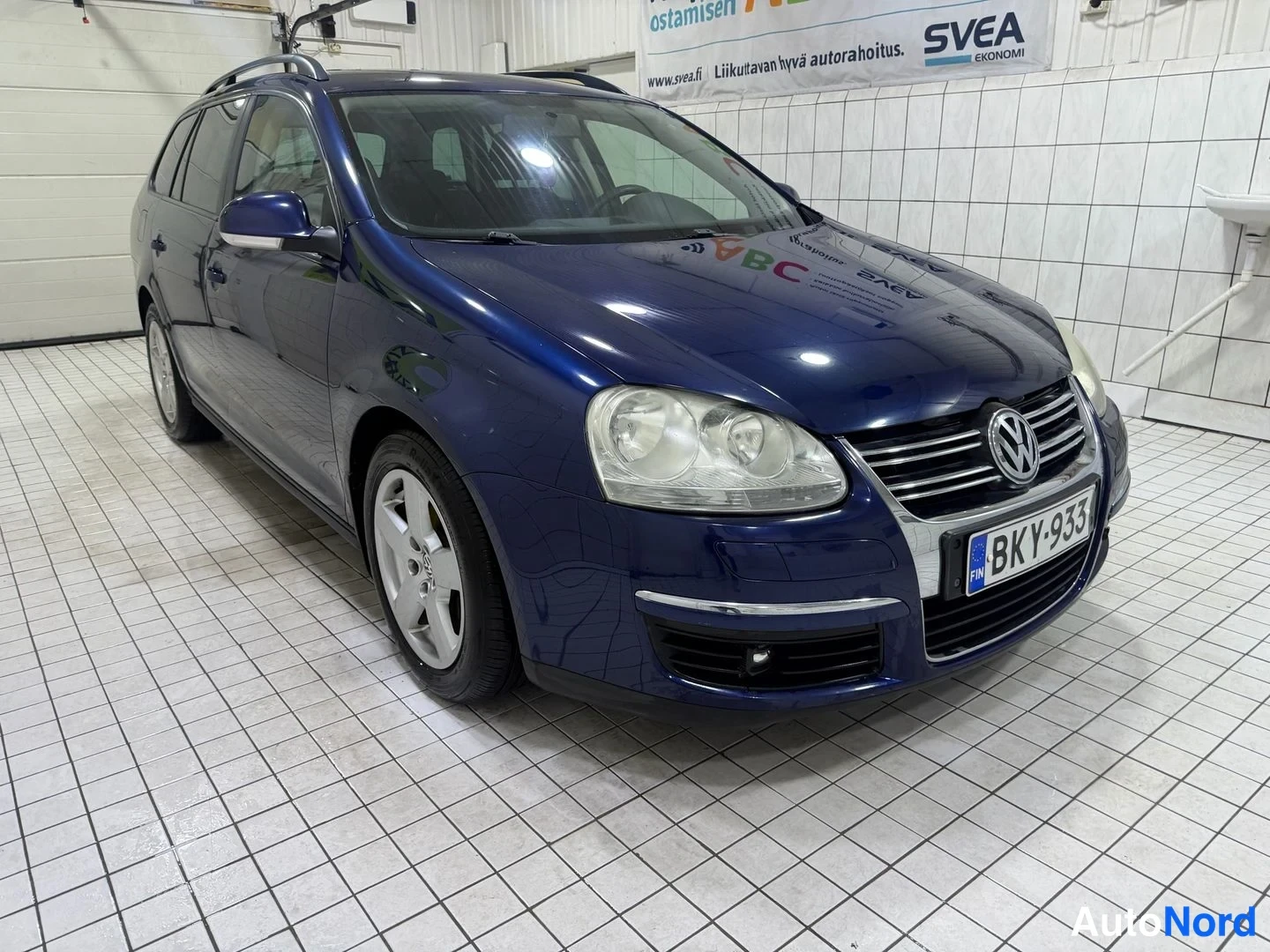 Etuviisto, nokka oikealle: Volkswagen Golf Variant -farmariauto edestä vasemmasta kulmasta valokuvattuna sisätiloissa; tummansininen koripinta, kattokaiteet ja rekisteritunnus näky