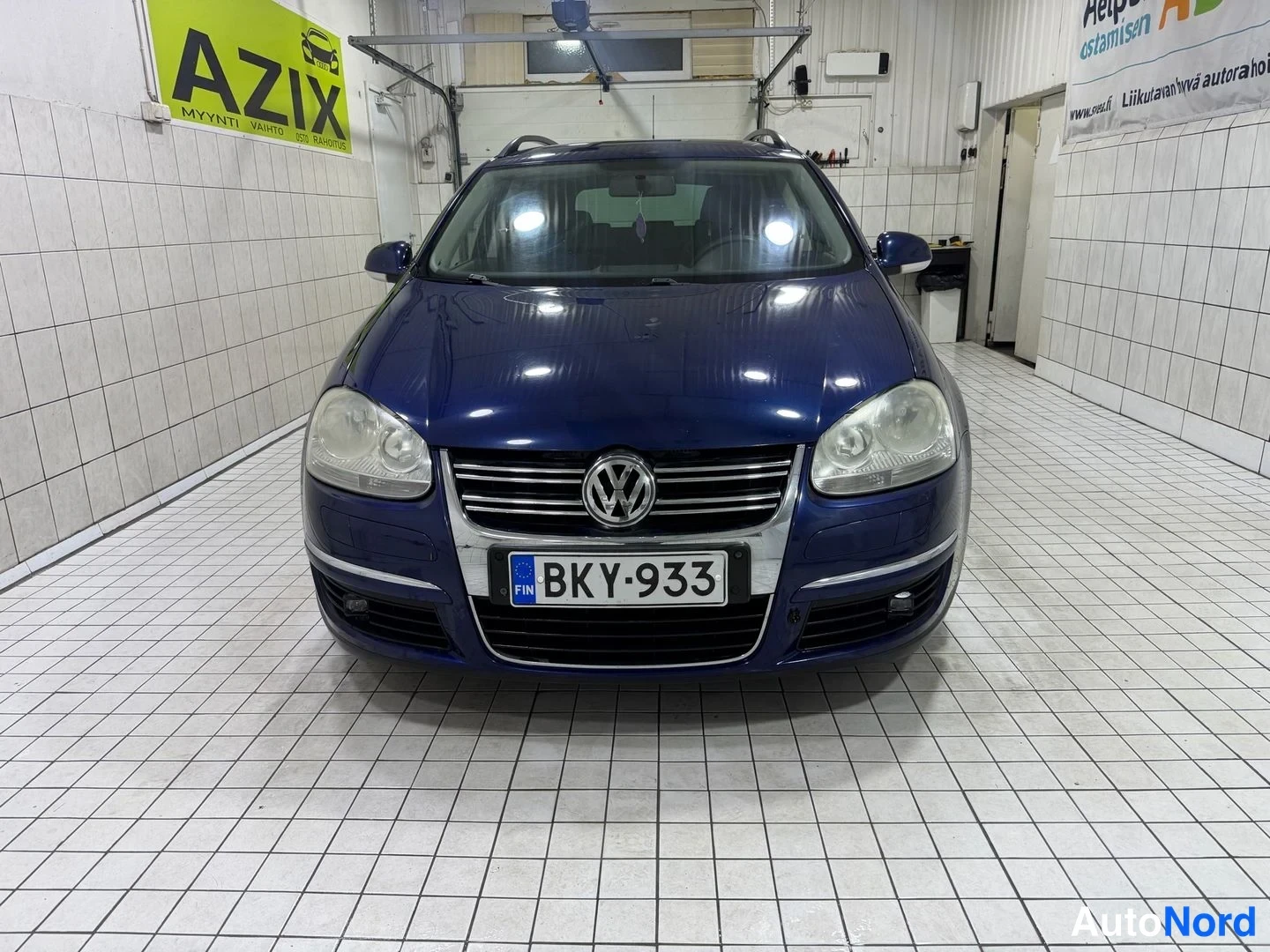 Edestä: Volkswagen Golf Variant -farmariauto suoraan edestäpäin valokuvattuna sisätiloissa; tummansininen koripinta, kattokaiteet ja rekisteritunnus näkyvät.