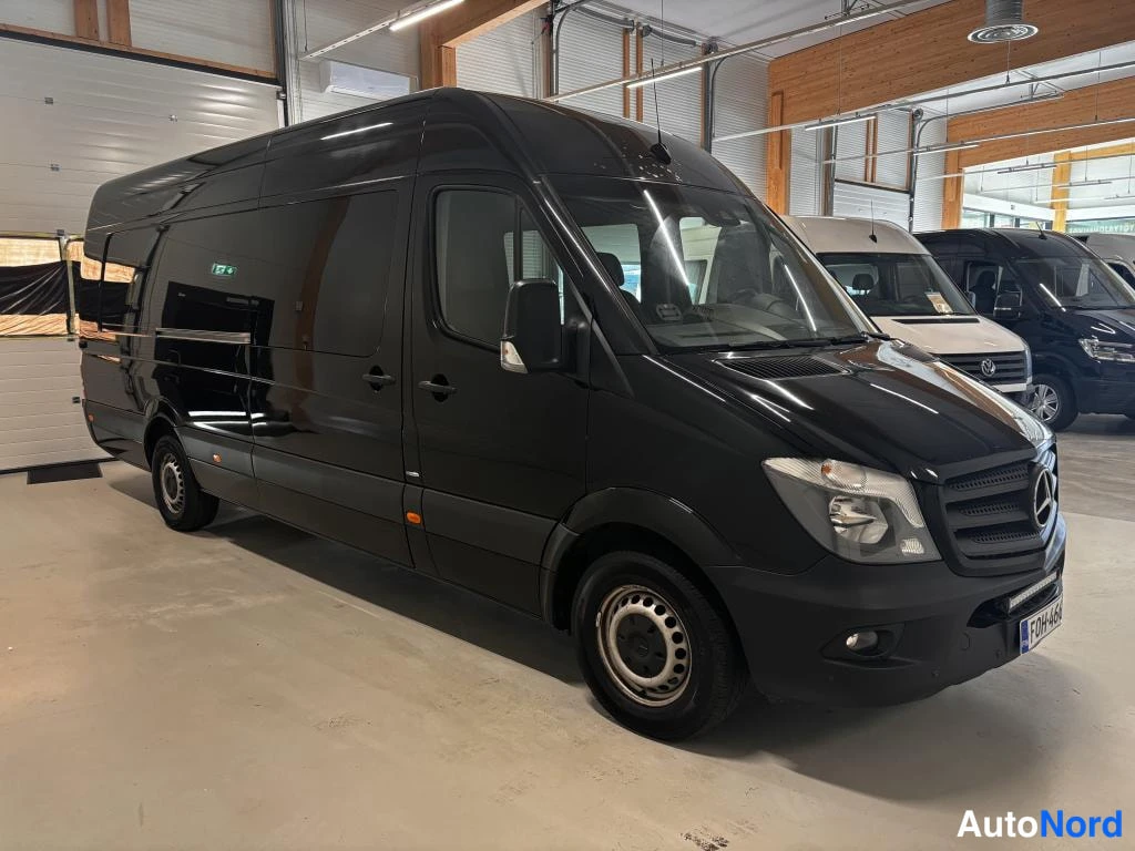 Etuviisto, nokka oikealle: Musta Mercedes-Benz Sprinter -pakettiauto kuvattu kolmivinkeltestä edestä oikealta hallissa, koko ajoneuvo näkyvissä.