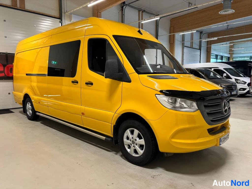 Etuviisto, nokka oikealle: Keltainen Mercedes-Benz Sprinter -pakettiauto etuviistöstä oikealta, pitkä korirakenne ja panssariverkko ikkunassa näkyvissä autotallissa.
