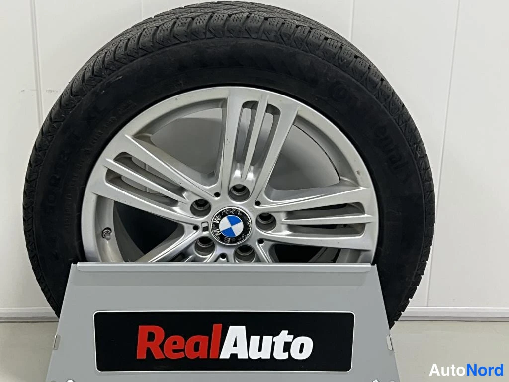 Sivulta: Rehellinen talvirengas BMW-merkki­vanteella, jossa on RealAuto-logolla varustettu tumma teline valkoisella taustalla. Rengasmerkinnät eivät ole täysin luettavis