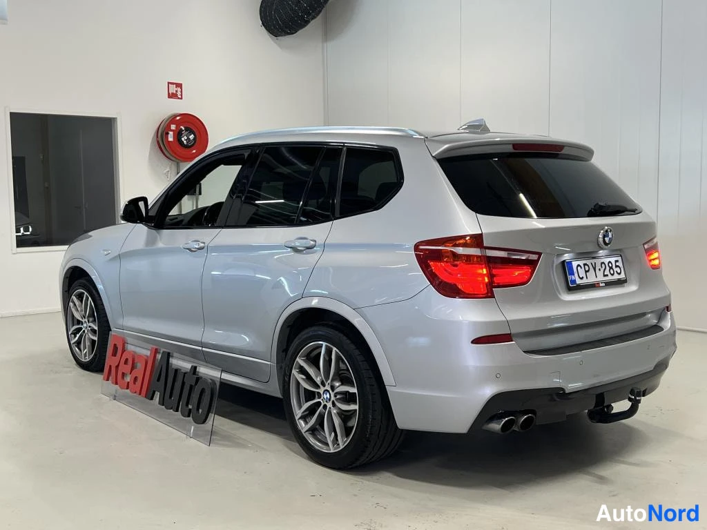 Takaviisto: Hopeanvärinen BMW X3 farmarimallinen takaa kulmasta kuvattuna, rekisteritunnus, takavaloissa kirkas valo, sisätilassa. Takapuskurissa huomataan vetokoukku ja ka