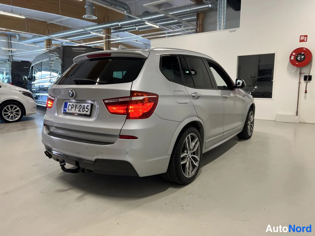 Takaviisto: Hopeanvärinen BMW X3 takaa kulmasta kuvattuna autotallissa, rekisteritunnus, takavaloissa kirkas valo, taustalla muita ajoneuvoja.