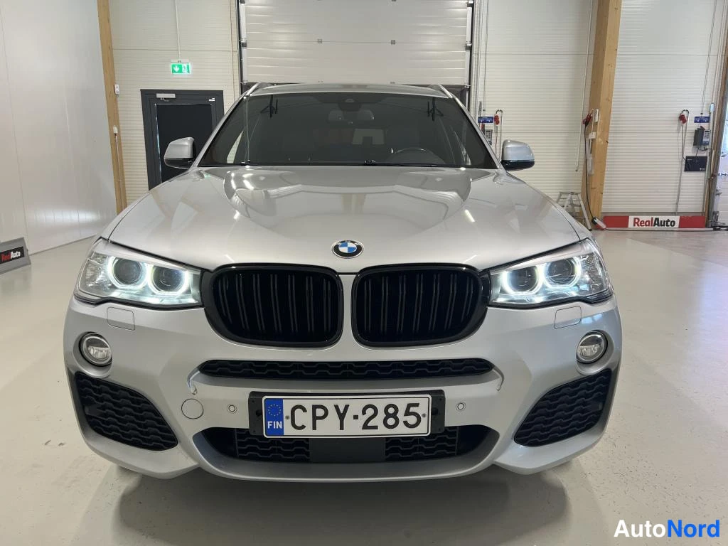 Edestä: BMW:n etunäkymä: valoisa etukolari, musta munuaisritilä, sumuvalot ja rekisterikilpi autotallissa.