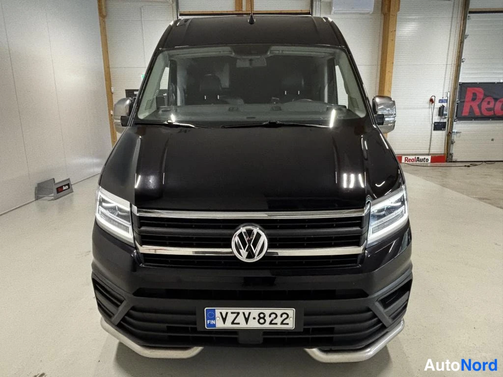 Edestä: Musta pakettiauto suoraan edestä kuvattuna hallissa; näkyvissä VW-merkki, ajovalot ja rekisterikilpi.