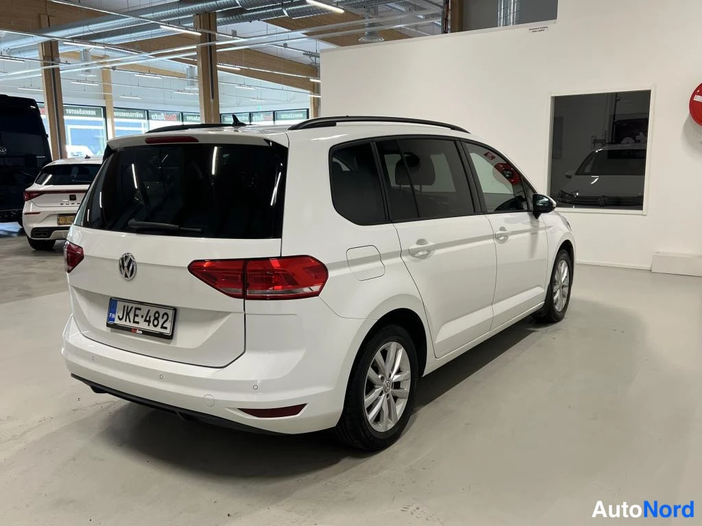 Takaviisto: Valkoinen farmarimallisesti muotoiltu tila-auto takaa ja oikealta kyljeltä kuvattuna autosalissa, kattokaiteet ja -rekisterikilpi näkyvät.