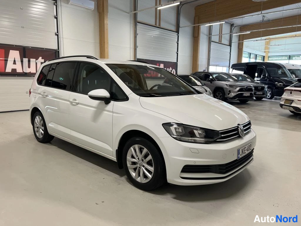 Etuviisto, nokka oikealle: Valkoinen tila-auto etukulmasta ja vasemmalta kyljeltä kuvattuna autosalissa, kattokaiteet ja kevytmetallivanteet näkyvät.