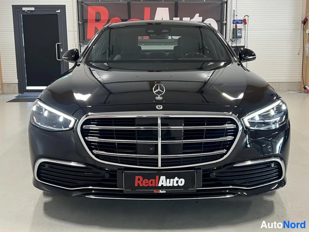 Edestä: Musta Mercedes-Benz S-luokan sedan suoraan edestä, kyynärvalot päällä ja RealAuto-kilpi etupuskurissa.