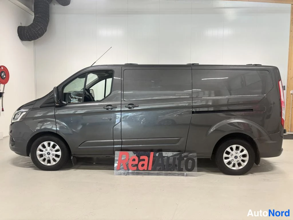 Etuviisto, nokka vasemmalle: Ford Transit Custom -pakettiauto sivulta vasemmalta, tummanharmaa, liukuovet ja hopeavanteet näkyvissä.
