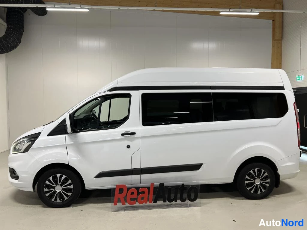 Etuviisto, nokka vasemmalle: Valkoinen Ford Transit Custom -pakettiauto lähes suoraan sivulta, kevyt etuviisto vasemmalta; vasen kylki, liukuova ja etuvalo näkyvät sisähallissa.