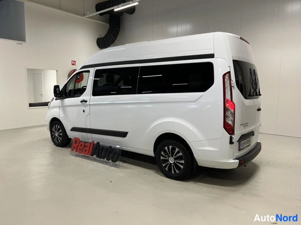 Takaviisto: Valkoinen Ford Transit Custom -pakettiauto kuvattuna takaviistosta oikealta sisähallissa; takavalot, takaluukku ja oikea kylki näkyvät.