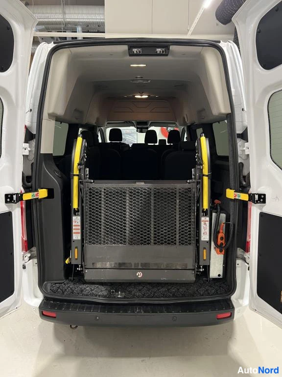 Takaa: Ford Transit Custom -pakettiauton tavaratila takaa avattuna; lattialla taitettu pyörätuolihissin musta lava ja molemmin puolin keltaiset kaiteet, takapenkki etu