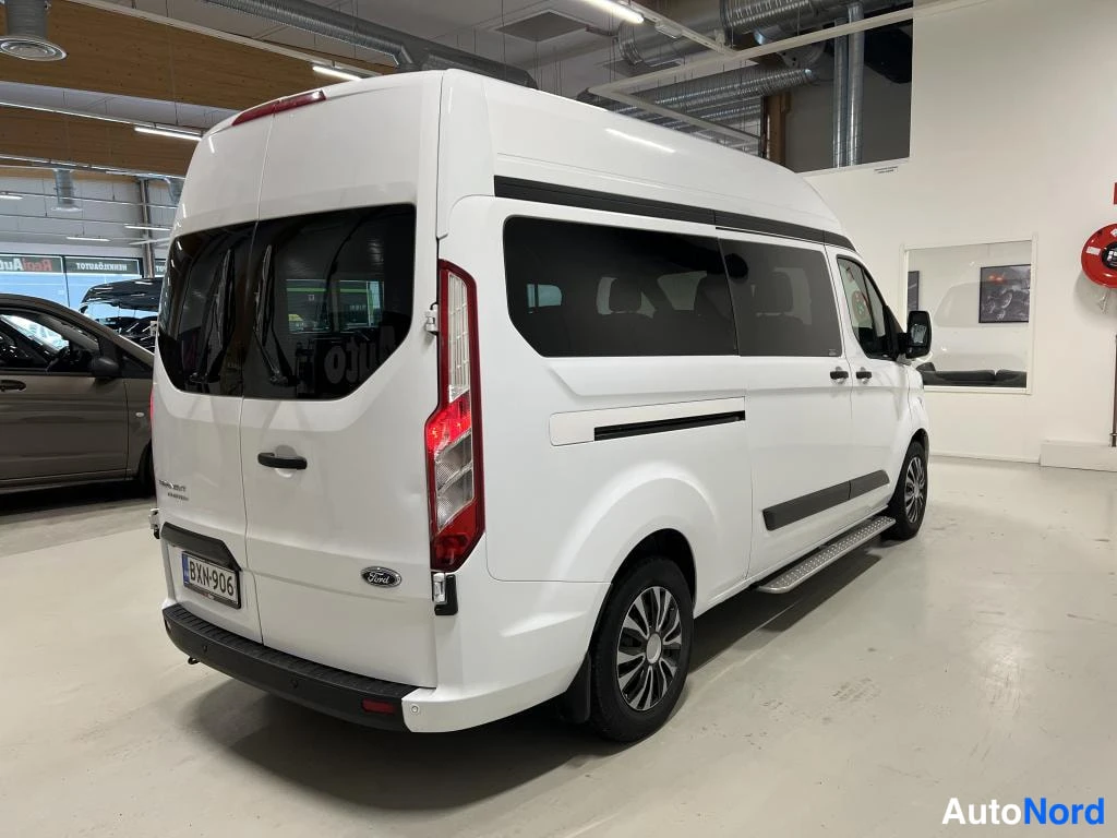 Takaviisto: Valkoinen Ford Transit Custom korkealla katolla takaa sivulta kuvattuna; rekisterikilpi, kromiset sivuaskelmat ja mustat vanteet, autotallissa sisätiloissa.